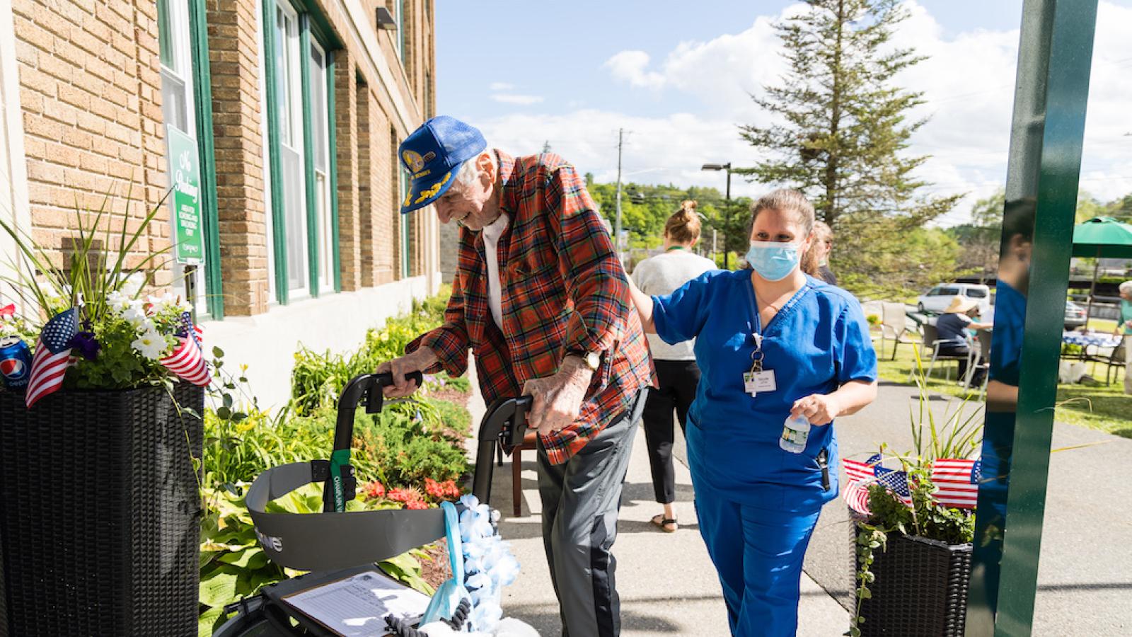 A resident using a walker entering our brick building. He is receiving help from a nurse.