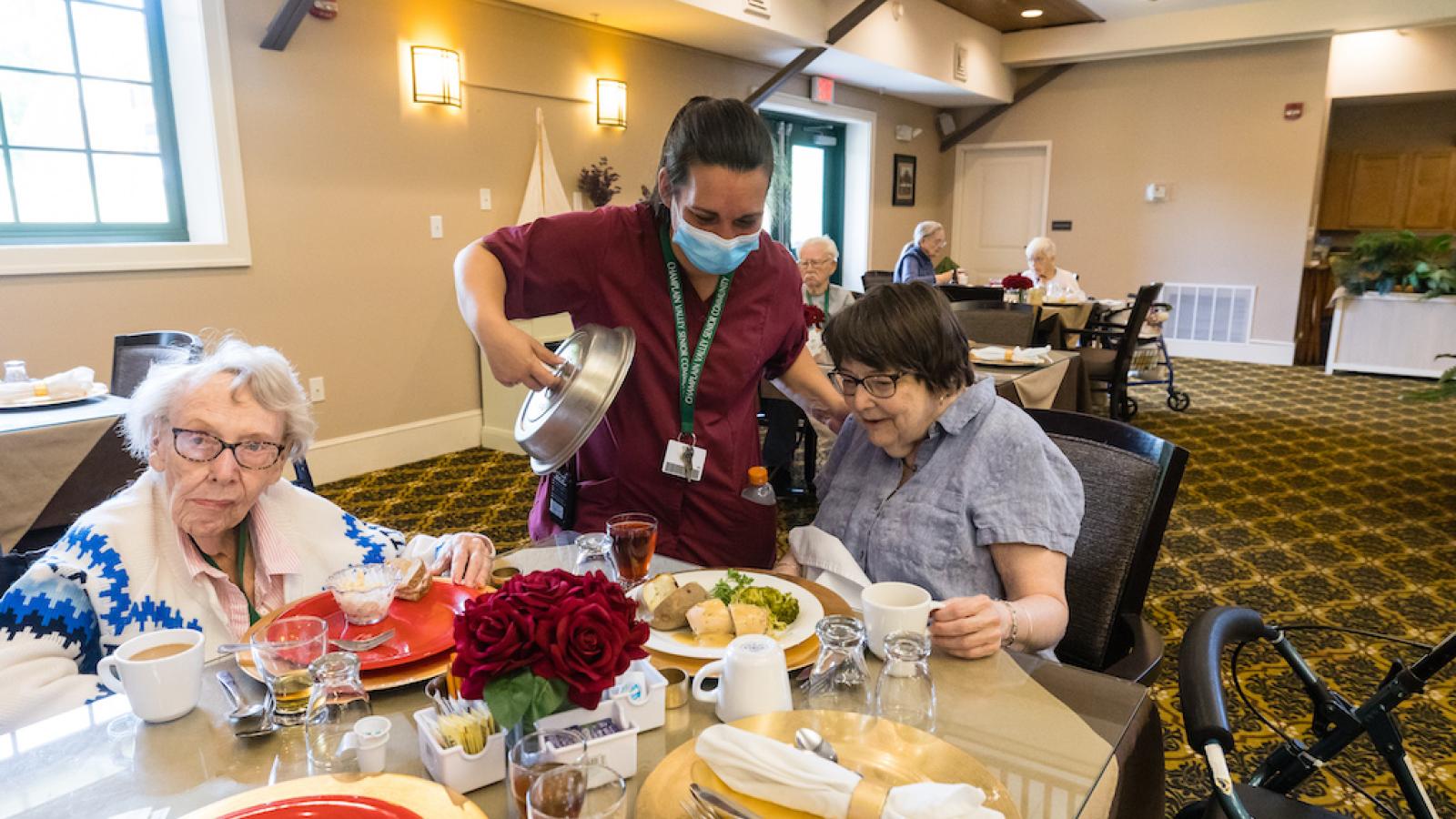 Two residents in the dining room looking at their meals, as a nurse unveils another plate.