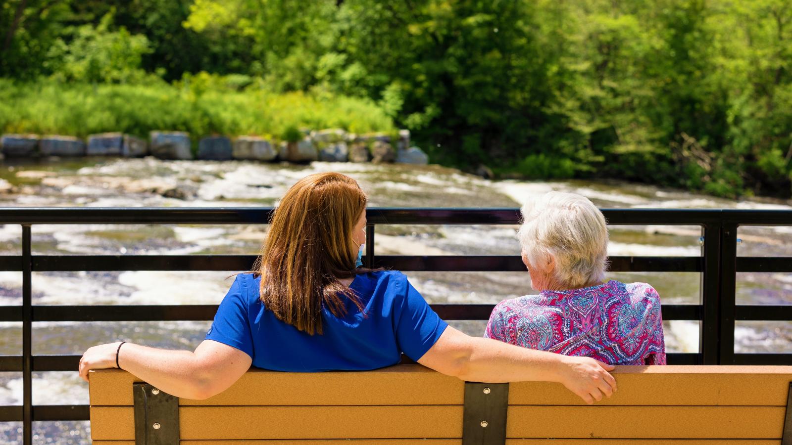 Staff and resident seated on bench watching river, backs to camera.